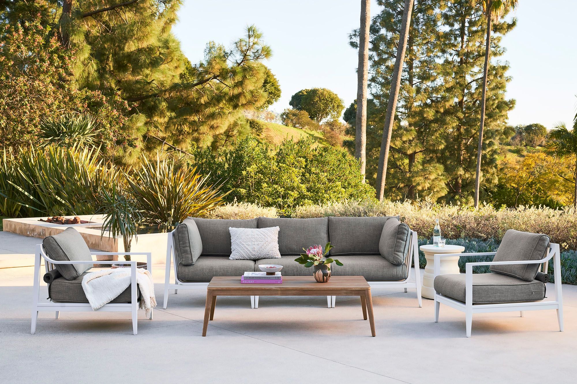 White aluminum 3 seat sofa in pebble gray with matching armchairs arranged outdoors with the tapered leg teak coffee table.